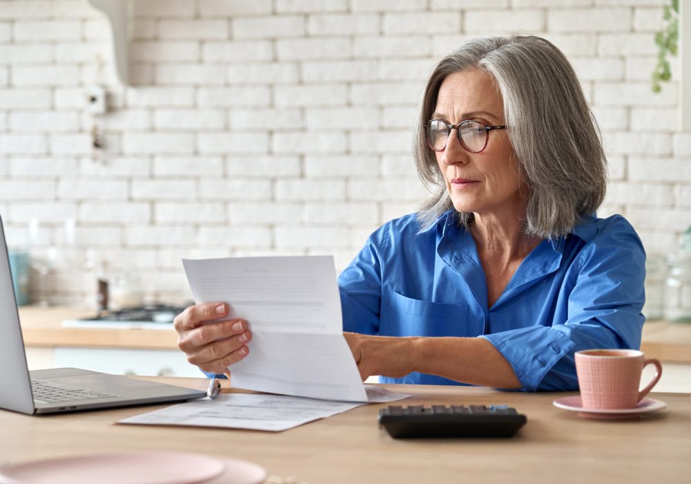 Adult senior 60s woman working at home at laptop. Serious middle aged woman at table holding document calculating bank loan payments, taxes, fees, retirement finances online with computer technologies